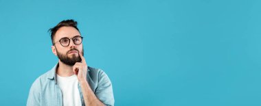 Sakallı ve gözlüklü bir adam derin düşüncelere dalmış başını kaldırırken çenesine dokunuyor. Sıradan bir gömlek giyiyor ve parlak mavi bir arka plana karşı duruyor. Rahatlamış bir ortam yaratıyor..