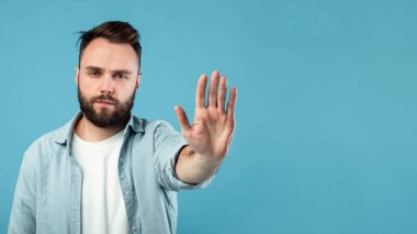 Sakallı ve kısa saçlı bir adam sağlam bir mavi arka plana karşı duruyor. Bir elini kaldırır, dikkat çekmek ya da durmak için işaret eder. Yüzündeki ifade ciddi ama sakin görünüyor, bir anlık duraksama yaratıyor..