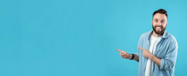 Sakallı ve dostça gülümseyen bir adam eliyle boş bir alana doğru hareket eder. Sıradan giyinmiş ve parlak mavi bir duvarın önünde duruyor..