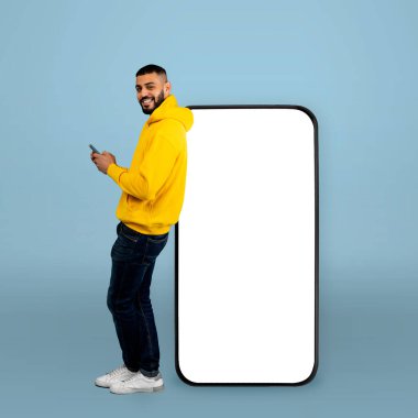 Sarı kapüşonlu bir adam boş bir ekranın yanında duruyor, akıllı telefonunu kullanırken gülümsüyor. Arka plan yumuşak bir mavidir, modern, canlı bir atmosfer yaratır..