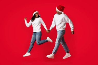 İki insan mutlu bir şekilde el ele tutuşuyor ve birlikte zıplıyorlar, ikisi de Noel Baba şapkaları ve rahat kazaklar giyiyor. Neşeli enerjileri ve parlak kıyafetleri bayram için mükemmel bir ortam yaratıyor..