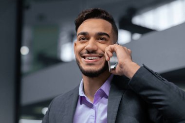 Takım elbiseli genç bir adam ofisin içinde telefonla konuşuyor. İletişim kurarken mutlu ve meşgul görünüyor. Doğal ışık pencereden içeri giriyor..