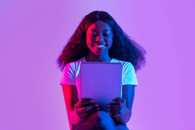 Odaklanmış genç siyah bir kadın dizüstü bilgisayarı üzerinde çalışıyor neon ışıklarla aydınlatılmış yaratıcı bir alanda. Modern bir uzaktan çalışma ortamı sunan bir video toplantısıyla meşgul görünüyor..