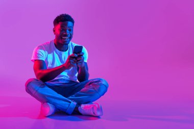 Bin yıllık Afro-Amerikan bir adam bir arkadaşıyla sohbet etmek için akıllı telefonunu kullanırken bacak bacak üstüne atmış, gülümsüyor. Sahne parlak neon renklerle aydınlanıyor, canlı bir atmosfer yaratıyor..