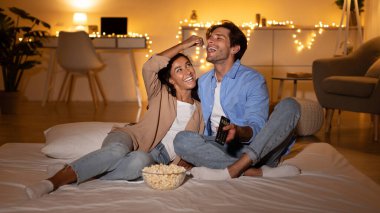 Mutlu çift yataklarında rahatça oturup patlamış mısırlarını paylaşıyor ve birbirlerini besliyorlar. Yumuşak ışıklandırma ve evlerinde rahat bir atmosferle çevrili bir film gecesinin tadını çıkarıyorlar..