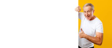 Bir adam parlak sarı bir arka plana karşı duruyor. O geniş bir şekilde gülümser ve bir eliyle baş parmağını kaldırırken diğer eliyle diğer elini işaret eder. Sahnenin neşeli bir havası var..