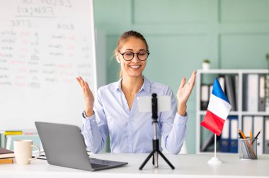 Neşeli bir bayan öğretmen öğrencileri uzak bir Fransızca dersine dizüstü bilgisayar ve akıllı telefon kullanarak çekiyor. Sahne, canlı bir öğrenme ortamı sunan, Fransız bayrağıyla aydınlık bir çalışma alanı..