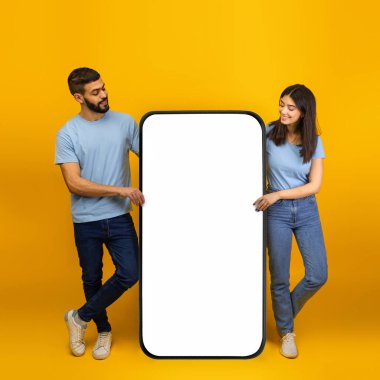 Milenyum Arap bir çift, büyük bir akıllı telefon modelinin önünde duruyor ve havalı bir mobil uygulamayı tanıtıyorlar..