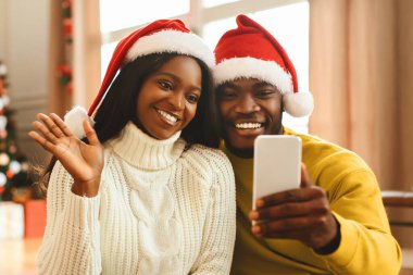 Bir çift birlikte oturuyor, ikisi de kırmızı Noel şapkası takıyor. Akıllı telefondan selfie çekerken gülümserler. Arka planda tatil süslemeleriyle şenlikli bir ortam var..