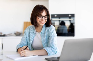 A woman with glasses is sitting at a kitchen table, smiling as she studies on her laptop while taking notes. The kitchen is bright and stylish, offering a calm atmosphere for learning.