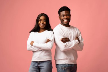 Kendine güvenen genç bir zenci erkek ve kadın sırt sırta verip kollarını kavuşturup kameraya mutlu bir şekilde gülümsüyor. Canlı pembe bir arka planda günlük beyaz kazaklar giyiyorlar..