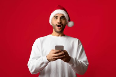 Noel Baba şapkası takan bir adam, Noel sezonunda canlı kırmızı bir arka planda akıllı telefonuna bakarken heyecan gösteriyor..