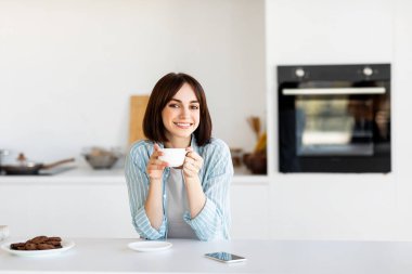 Genç bir kadın mutfak tezgahında oturuyor, elinde bir fincan kahve tutarken gülümsüyor. Kurabiyeler yakına yerleştiriliyor ve telefon tezgahın üzerinde. Parlak sabah ışığı odayı dolduruyor..