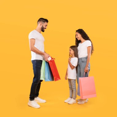 Gülümseyen bir aile alışveriş yaparken neşeli bir anı paylaşır. Baba renkli alışveriş torbaları tutuyor, ve kız heyecanlı görünüyor, anne mutlu bir şekilde izliyor, sıcak bir atmosfer yaratıyor..