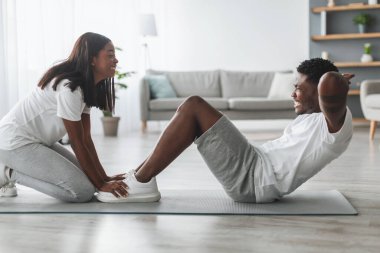 Evde karın kasları üzerinde çalışan mutlu, genç, siyah bir çift mekik çeken, mekik çeken, profil manzaralı, oturma odasının içini gören bir kadın. Fitness, tecrit sırasında egzersiz