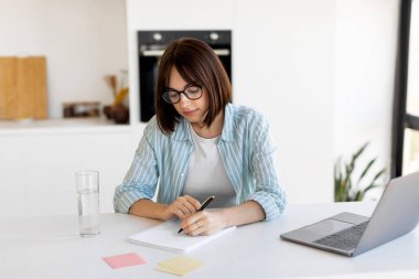 Gözlüklü bir kadın masasında otururken bir deftere notlar yazıyor. O bir bardak su ve bir dizüstü bilgisayarla çevrilidir. Doğal ışık odayı dolduruyor..
