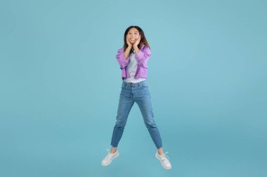 Gündelik giysiler giyen genç bir kadın heyecanla havaya zıplıyor, parlak bir şekilde gülümsüyor. Canlı, mavi bir arkaplanda, eğlenceli ve canlı bir atmosfer yaratıyor..