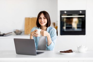 A young woman sits at a kitchen counter, smiling as she holds a cup of coffee. A laptop is open in front of her, along with a plate of cookies and a teapot, creating a cozy work atmosphere.