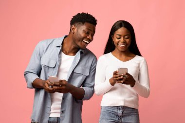 A happy African American couple in casual outfits play mobile games on their new smartphones. They are smiling and enjoying each others company against a pink background, creating a fun atmosphere.