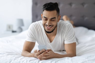 A happy young Arab man lies comfortably in bed, smiling as he messages friends or browses the internet on his smartphone. He enjoys his downtime in the cozy bedroom.
