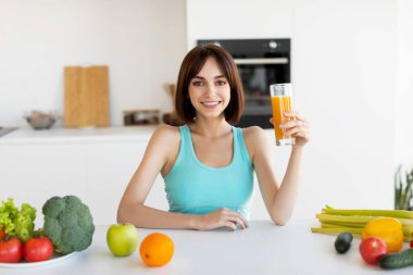 Neşeli bir kadın, taze meyve ve sebzelerle çevrili temiz bir mutfak masasında otururken bir bardak portakal suyu tutuyor. Ilık aydınlatma sağlıklı atmosferi güçlendirir..