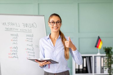 Neşeli bir bayan öğretmen Almanca öğretirken geniş bir şekilde gülümsüyor. Parlak bir sınıfta arka planda bir Alman bayrağı ve beyaz tahtası olan bir not defteri tutuyor..