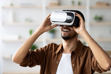 Genç bir adam sanal gerçeklik deneyimi yaşıyor, kulaklık takıyor ve gülümsüyor. Parlak ve modern bir odada. Dekoru az. Entrikacı etkinliğinin tadını çıkarıyor..