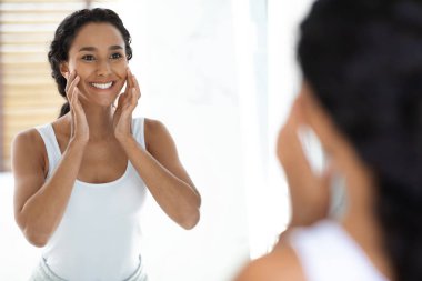 Siyah saçlı bir kadın aynanın önünde duruyor, yüzüne nazikçe dokunurken gülümsüyor. Parlak bir banyoda cilt bakımından hoşlanıyor gibi görünüyor..