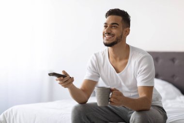 Genç Arap adam televizyon izlerken gülümseyerek yatak odasında dinleniyor. Kahve içiyor ve uzaktan kumandayla kanal değiştiriyor. Sıcak atmosfer boş zamanlarını artırıyor..
