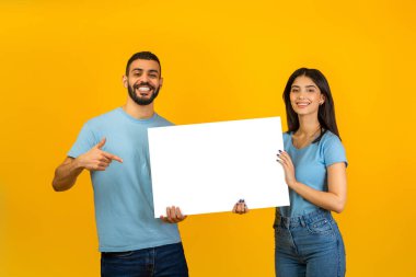 Neşeli Arap çift birlikte poz veriyor, reklam için beyaz bir afiş tutuyorlar. Adam, parlak sarı bir arkaplanda dururken mekanı işaret eder..