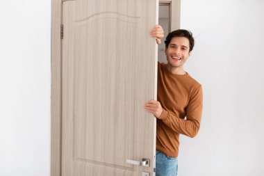 Neşeli genç bir adam modern bir dairenin kapısında duruyor, kapıyı aralık tutarken gülümsüyor ve dışarı bakıyor. Misafirperver ve dost canlısı görünüyor, bir ziyaretçiyi karşılamaya hazır..