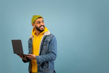 Bir adam açık mavi bir arkaplanda dizüstü bilgisayarını tutarken gülümsüyor. Kot ceketinin altına yeşil bir bere ve sarı kapüşonlu bir sweatshirt giyiyor. Nişanlı ve mutlu görünüyor..