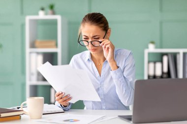 Kadın ofis çalışanı masasında oturup evrak işlerine odaklanmış. Gözlüklerini ayarlıyor, ofisindeki belgeleri okumaya çalışıyor. Çevre düzenli ve profesyonel..