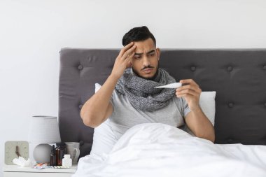 Genç bir Arap adam yatakta, atkıya sarılmış, termometre tutarken hastalık belirtileri gösteriyor. Vücut ısısını ölçerken hayal kırıklığına uğramış görünüyor. Bu da grip belirtilerini gösteriyor..