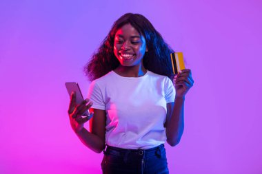 Coşkulu bir Afrikalı Amerikalı genç kadın akıllı telefonunu internet üzerinden ticaret yapmak ya da kumar oynamak için kullanıyor. Elinde bir kredi kartı tutarken gülümsüyor, etrafındaki neon ışığın tadını çıkarıyor..