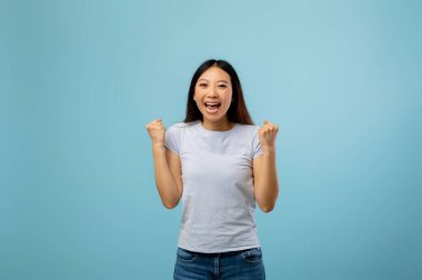 Genç bir kadın sevinçle yumruğunu açık mavi bir arka plana kaldırarak heyecanını ifade eder. Kocaman bir gülümsemesi var ve sıradan giyinmiş, bir anlık mutluluk gösteriyor..