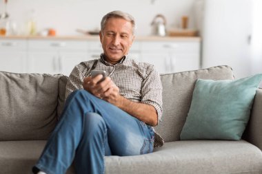 Günlük kıyafetli bir adam akıllı telefonuna bakarken bir kanepede oturuyor. Rahat ve hareketleriyle meşgul görünüyor. Rahat bir oturma odasında..