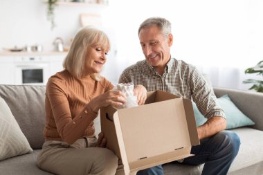 Yaşlı bir çift rahat bir kanepede oturuyor, bir hediyeyi açarken gülümsüyor. Güneş ışığı pencereden akar ve bu paylaşılan anın tadını çıkarırken sıcak, davetkar bir atmosfer yaratır..