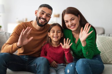 Üç kişilik neşeli bir aile evdeki kanepede oturup kameraya el sallıyor ve gülümsüyor..