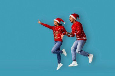 Renkli Noel süveteri ve Noel Baba şapkası giymiş bir çift kutlama için atlıyor. Tatil sezonunda neşe ve coşkuyu ifade ederler. Parlak mavi arka plan şenlik havasını arttırır..