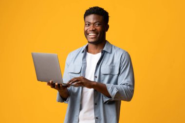 Beyaz t-shirt ve kot gömlek giyen yakışıklı siyahi adam sarı stüdyo arka planında modern laptop kullanıyor, bilgisayar klavyesinde daktilo ediyor, çöpçatan sitesinde kadınlarla sohbet ediyor, boşluğu kopyalıyor.