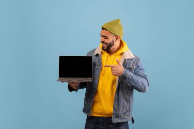 Sarı kapüşonlu ve kot ceketli bir adam elinde tuttuğu dizüstü bilgisayarı işaret ederken gülümsüyor. Arka plan açık mavi, neşeli bir atmosfer yaratıyor..