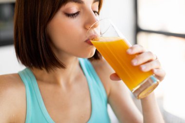 Kısa kahverengi saçlı genç bir kadın bir bardak portakal suyu içer. Açık mavi bir bluz giyiyor ve güneşli bir mutfakta hafif ışıklandırmayla oturuyor..