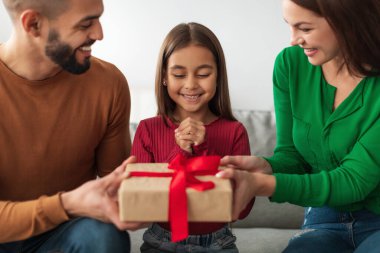 Küçük bir kız kanepeye oturmuş, ailesi ona bir hediye verirken heyecanla gülümsüyor. Aile, mutlu bir şekilde doğum gününü rahat evlerinde kutluyor..