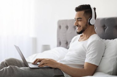 Genç Arap adam yatakta dizüstü bilgisayarını kullanırken gülümsüyor ve odaklanıyor. Kablosuz kulaklık takıyor ve uzak bir iş oturumunda rahat ortamının tadını çıkarıyor..