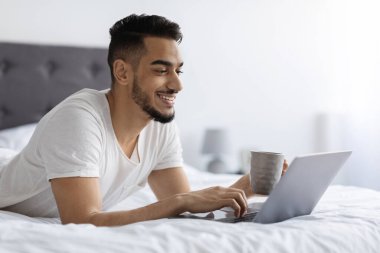 Yakışıklı bir Arap, dizüstü bilgisayarını kullanarak ve kahvesini yudumlayarak rahatça yatakta uzanıyor. İnternette gezinirken mutlu görünüyor, yatak odasında biraz boş vakit geçiriyor..