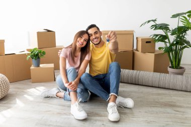 Mutlu çift yeni evlerinin zemininde oturup anahtarlarını gösteriyor. Etrafları taşınan kutularla ve kapalı alandaki bitkilerle çevrili, neşeli bir anı paylaşıyorlar..