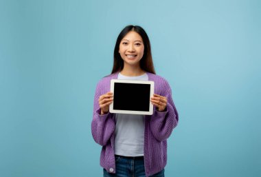 Neşeli genç bir kadın elinde tabletle açık mavi bir arkaplanda duruyor. O, doğrudan izleyiciye bakarken parlak bir şekilde gülümser..