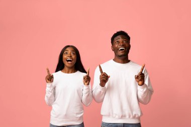 Genç Afro-Amerikalı erkek ve kadın sevinçle mesaj ve reklamlar için uygun boş bir alanı işaret ediyorlar. Beyaz kazaklar giyip pembe bir stüdyo arkaplanının önünde gülümsüyorlar..
