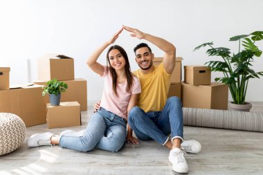 Mutlu bir çift, karton kutular ve ev bitkileriyle çevrili yeni oturma odalarının zemininde oturuyor. Elleriyle bir kalp şekli oluştururlar, aşkı ve yeni başlangıçları sembolize ederler..